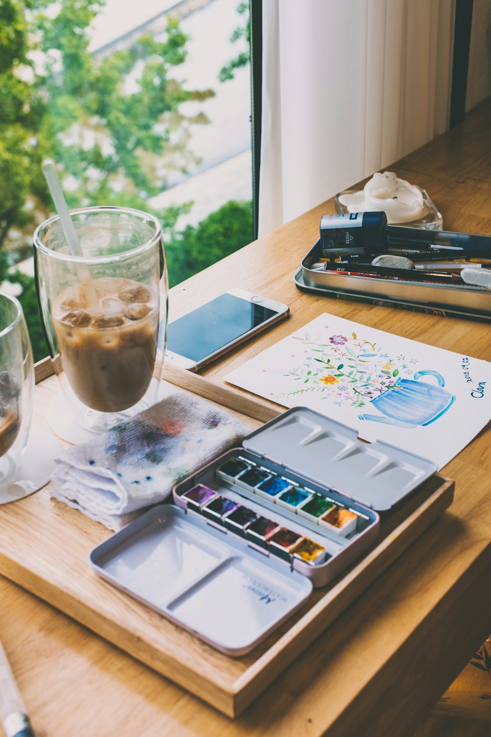 Art supplies and iced coffee on a wooden table workspace.