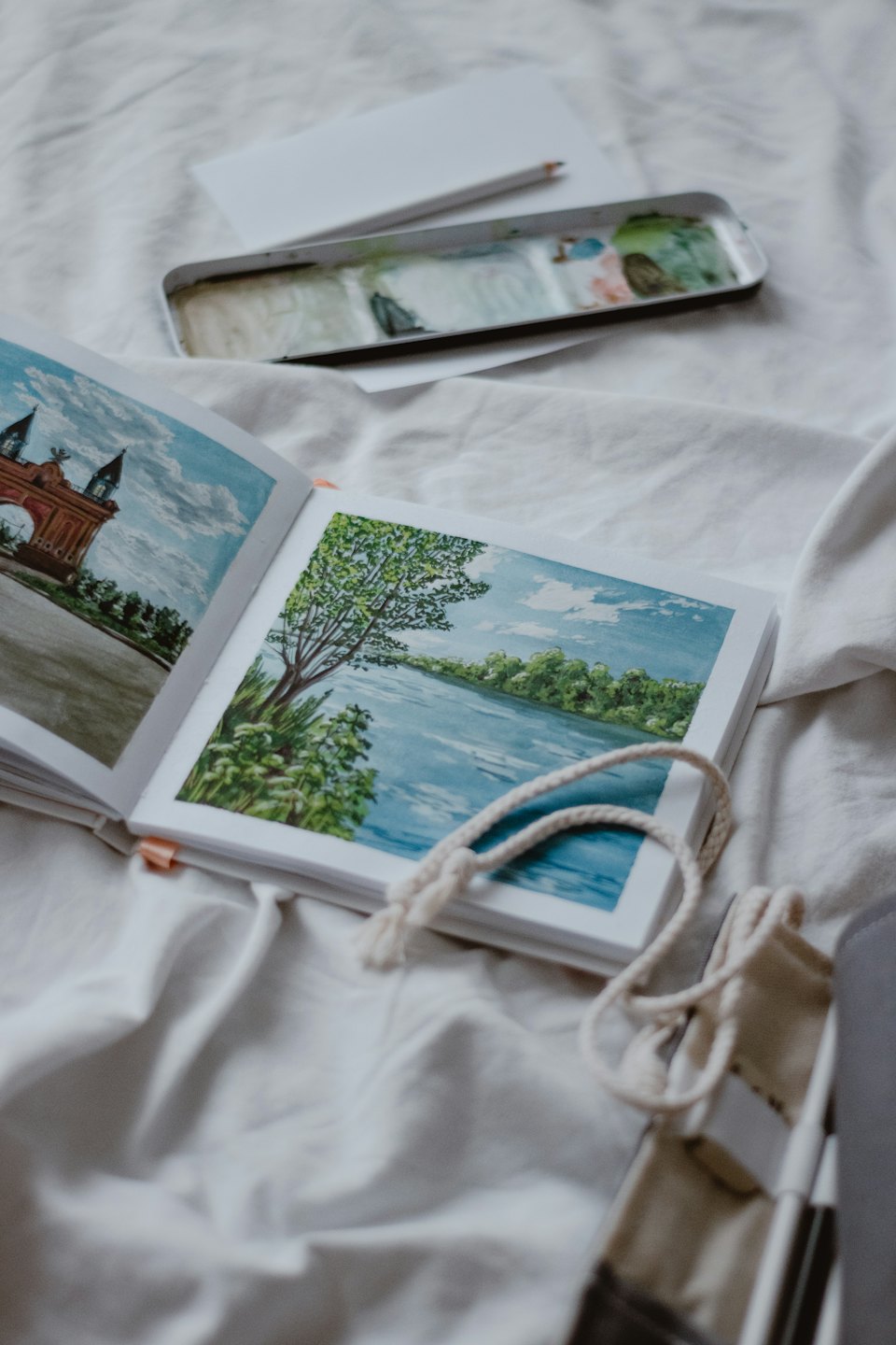 Open sketchbook displays watercolor landscape paintings on rumpled white fabric.