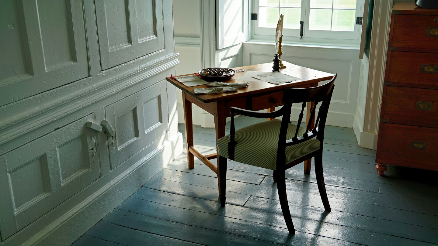 A wooden writing desk with papers and quill pen in a sunlit room.