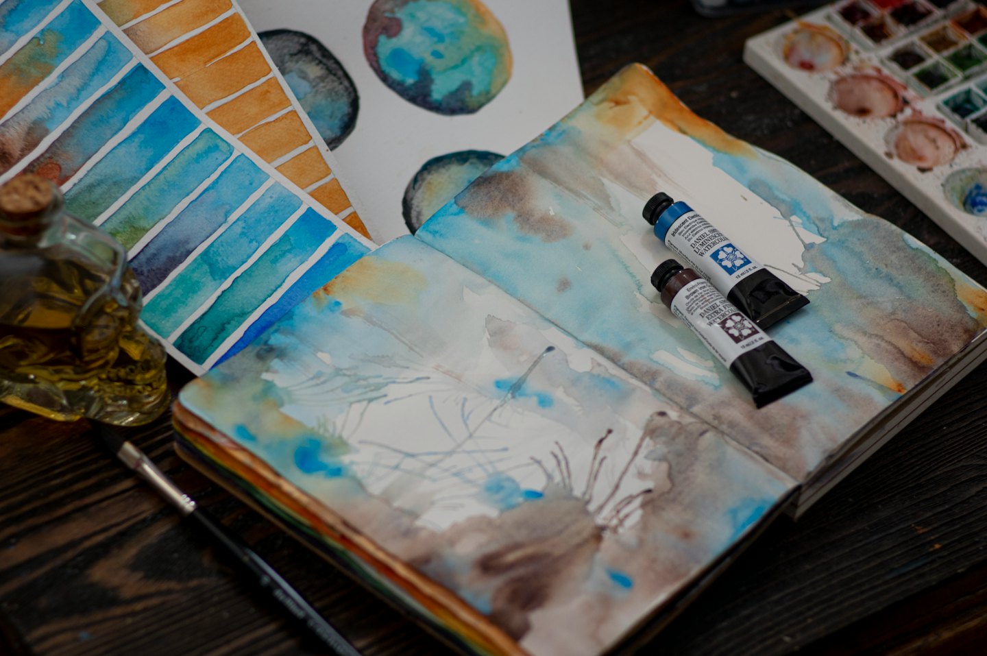 Artist's workspace with sketchbook, paints, and brushes on a wooden table.